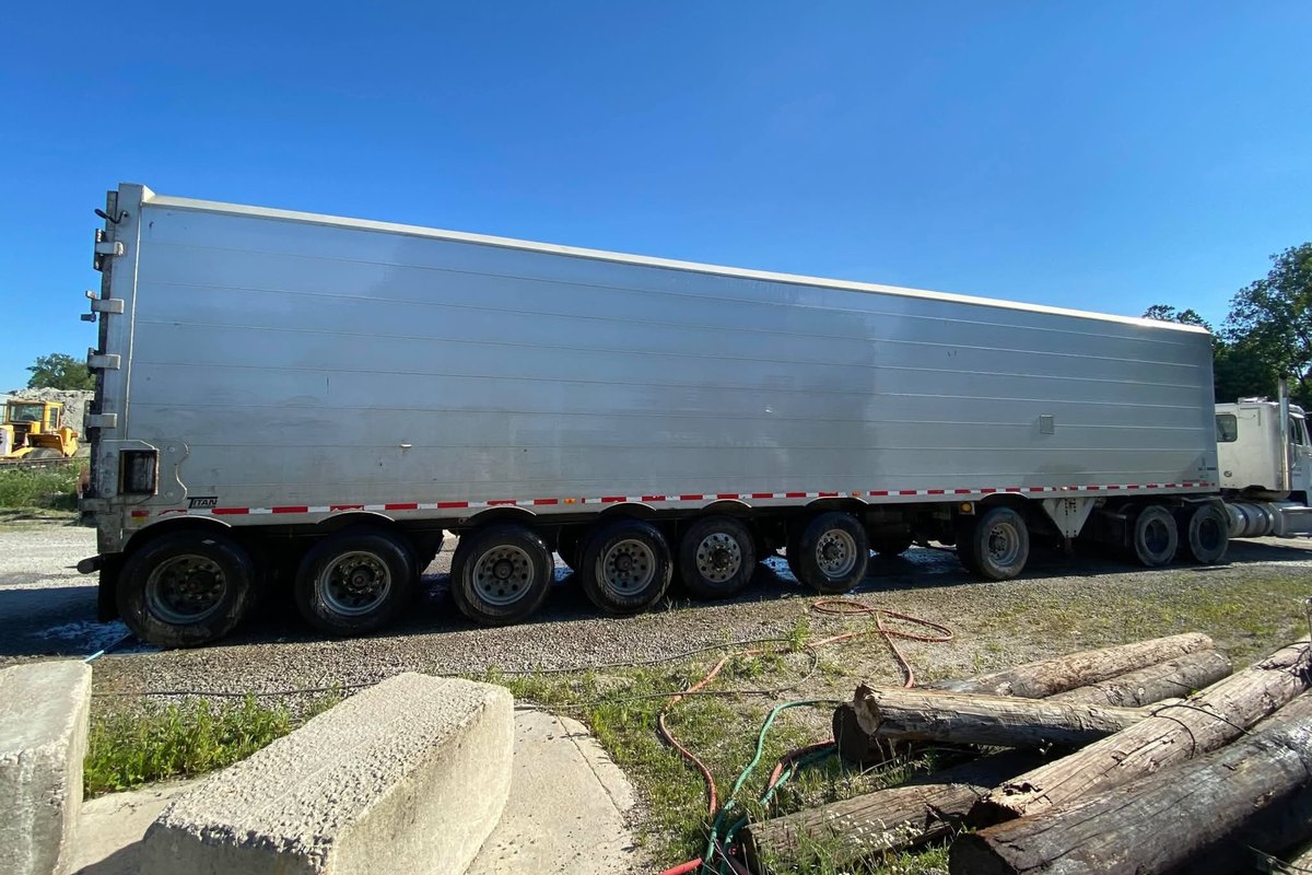 Long silver aluminum trailer on gravel with bright even panels, logs and concrete blocks in front.