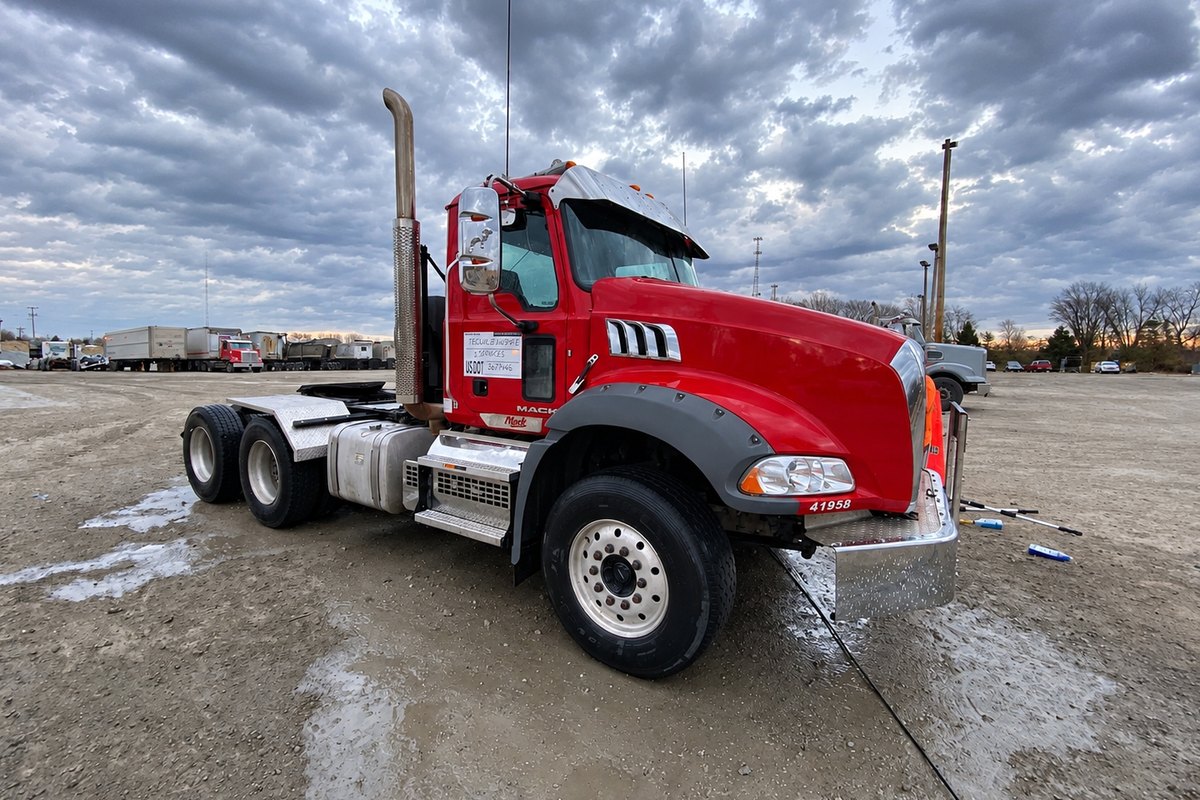 Clean red Mack tractor with reflective wheels, a silver fuel tank and steps on a wet gravel lot.